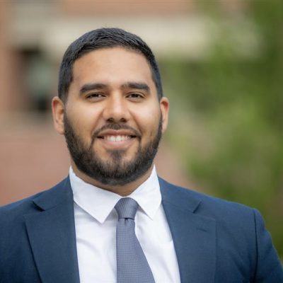 Man with short black hair and a short black beard, smiling and wearing a dark blue suit jacket, white shirt, and light blue tie in front of a brick building. 