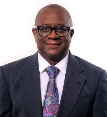 Black male wearing a jacket and tie. He has a smile on his face, he wears black glasses, and has very short hair