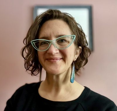 A woman wearing glasses and earrings stands in front of a vibrant pink wall.
