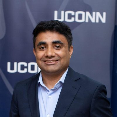 A man in a suit and tie stands confidently with a smile in front of a UConn sign, showcasing school pride and professionalism.