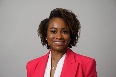 A woman wearing a pink blazer over a white shirt, standing confidently with a neutral background.
